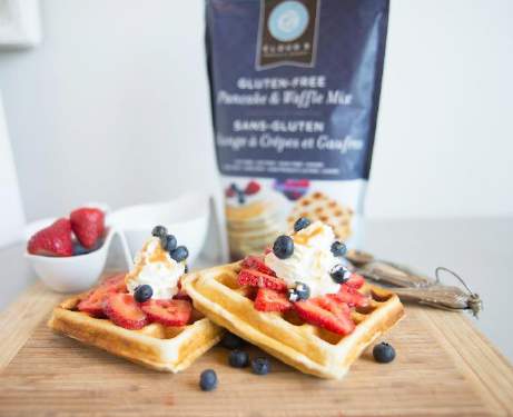 Two waffles covered in berries and cream sit on a wooden chopping board in front of a packet of Cloud 9 Pancake and Waffle mix.