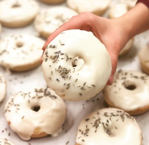A selection of donuts covered in white icing sit on a white countertop. A hand holds up one of the donuts to the camera.
